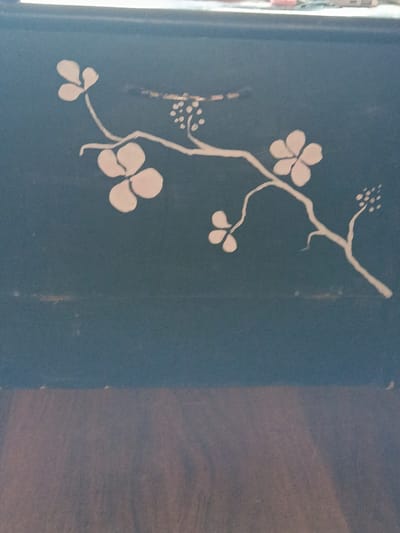 The image shows a close-up view of a dark blue surface with a white floral design. The design depicts a branch with several flowers and buds. The flowers have multiple rounded petals, and the buds are clustered together. The branch is thin and winding, with smaller twigs extending from it. The background is uniformly dark blue, with some slight texture visible. The bottom portion of the image shows a blurred area of brown, which appears to be a wooden surface. There are no people or actions depicted in the image. The location in the city of Dachne, Ukraine is not discernible from the image itself.