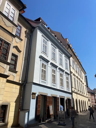 The image shows a street scene in Bratislava, Slovakia, with a narrow cobblestone street flanked by historical buildings. The buildings are multi-story with varied architectural styles and colors, including yellow, light blue, and cream. The facades feature multiple windows, some with ornate frames and shutters. The sky is clear and blue. On the right side of the street, there is a banner hanging from a building that reads "legendary night club". Several people are visible walking along the street. One woman is walking towards the camera carrying a bag, and another person is walking away from the camera in the background. A man in a blue shirt is visible standing inside one of the buildings. There are signs and displays outside some of the establishments. The scene captures a sunny day in a European city.