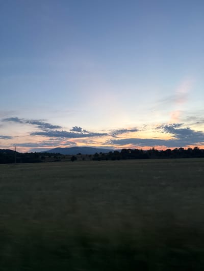 A wide-angle, low-angle shot captures a dusk scene over a field in Slokoshtitsa, Bulgaria. The sky transitions from a pale blue at the top to a warm blend of pink, orange, and yellow near the horizon. Scattered, wispy clouds are present in the mid-sky. A dark, undulating mountain range forms the backdrop, silhouetted against the colorful sky. In the middle ground, a line of trees with some houses nestled within them runs horizontally across the frame. To the left, a solitary utility pole with wires stretches upwards. The foreground is dominated by a dark, out-of-focus field, suggesting the photograph was taken from a moving vehicle.
