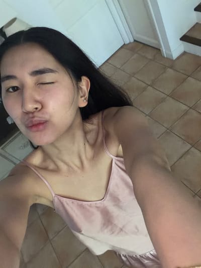 A fair-skinned young woman with long, dark hair is posing for a selfie. She is wearing a light pink satin camisole and is winking with her left eye. She has a piercing in her left ear. The background is comprised of light brown tiled flooring and a portion of a white staircase. The location in Grimaud, France, is unknown, as there are no identifying landmarks or features visible in the image.