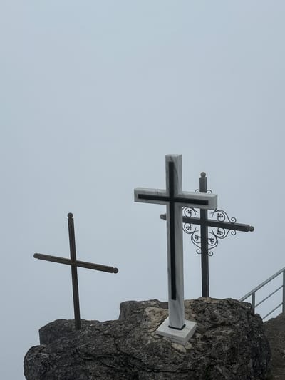 The media file shows three crosses mounted on a rocky outcrop. The crosses vary in size and material. The largest cross in the foreground appears to be made of white marble with black accents. Behind it, a smaller, black metal cross with ornate scrollwork is visible. Further to the right and lower down, another simple black metal cross is embedded in the rock. The scene is set against a foggy or overcast sky, suggesting a cool or damp weather condition. The lighting is diffused, indicating it could be morning or late afternoon, or simply a heavily overcast day. The rocky formation suggests an outdoor, elevated location, possibly a mountain or a cliff face. There are no people visible, and no text is present in the image. The overall mood is somber and reflective, perhaps evoking a sense of remembrance or spirituality due to the presence of the crosses. The location context provided is Dzirovani, Georgia.