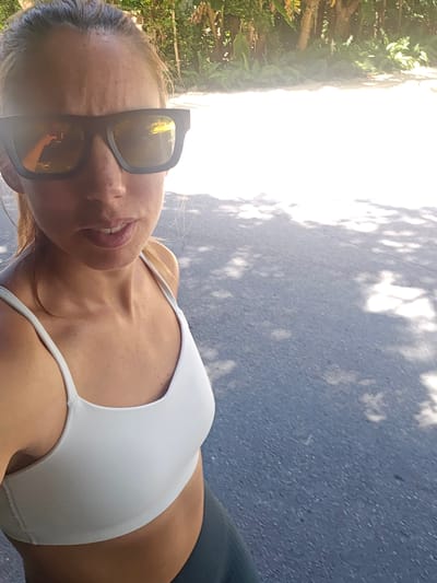 A woman, wearing a white sports bra and black sunglasses, takes a selfie outdoors. She has light brown hair and fair skin, with freckles visible on her décolletage. Her mouth is slightly open in a smile. The background shows a paved surface with dappled sunlight and shadows, suggesting an outdoor location like a park or sidewalk. To the right, lush green foliage and trees are visible, illuminated by bright sunlight, hinting at a warm, sunny day. The time of day appears to be daytime, possibly late afternoon, given the angle of the sun. The setting is likely Islamorada, USA, as indicated by the provided location context, which is known for its tropical climate and abundant greenery. There is no visible text in the image.