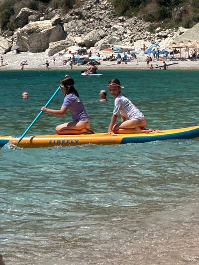 The media file depicts a lively beach scene at Mirište, Montenegro, bathed in bright sunlight, suggesting a midday time of day with clear weather. The primary subjects are several people enjoying a recreational activity in the water. In the foreground, two young girls are on a yellow paddleboard. One girl, wearing a colorful rash guard and pink sunglasses, is kneeling and looking towards the camera with a smile. The other girl, in a purple rash guard, is also kneeling on the paddleboard and holding a paddle, appearing focused on navigating the water.
The background reveals a bustling beach with numerous people relaxing on the sand, some under colorful umbrellas, and others wading or swimming in the turquoise water. The beach is backed by a rocky, vegetated hillside, characteristic of the Montenegrin coastline.
The activity taking place is paddleboarding and general beach recreation. The overall atmosphere appears joyful and energetic, with individuals engaging in water sports and enjoying a day at the beach.
Visible text on the paddleboard reads "FIREFLY". The scene is vibrant and full of life, capturing the essence of a summer day at a popular coastal destination. The water is clear and shimmering, reflecting the bright sunlight.