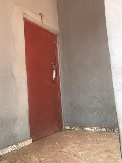 This forensic image captures an interior space within a building in Egboje, Nigeria. The scene is dominated by a reddish-brown, possibly metal, door positioned slightly left of center, set into a rough-textured, light grey concrete wall. A small, light-colored wooden crucifix is affixed to the wall directly above the door frame. The door is equipped with a silver-toned handle and latch assembly, and its lower portion exhibits a layer of accumulated dust or dirt. The floor is covered with patterned tiles in shades of tan and brown with dark, swirling lines, and is significantly soiled with numerous small, dark debris particles. A skirting board, composed of patterned white, grey, and black tiles, runs along the base of the walls. The wall to the right of the door is also light grey, rough concrete, showing minor surface imperfections and discoloration. No individuals, actions, or interactions are present in the static environment.