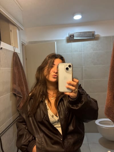 A selfie in a bathroom. A woman with long brown hair and a brown leather jacket holds up a phone to capture the image in a bathroom mirror. Bathroom fixtures like the toilet, towel rack with a brown towel, a light fixture, and a radiator are visible in the background. The bathroom is tiled in light gray and white. The woman wears a ring and a necklace. She is posing for the picture.