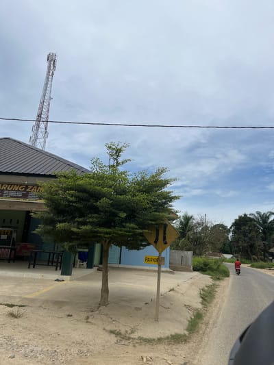 This is a street-level, medium-shot, eye-level photograph taken on a slightly overcast day in Benuaraja, Indonesia. In the foreground, a dirt road curves to the right, with a portion of a dark vehicle visible in the lower right corner. A lone individual is riding a motorcycle away from the viewer on the road. To the left of the road, there is a sandy area with sparse vegetation and a small, slender tree with dense green foliage. A diamond-shaped yellow sign with black markings is partially visible behind the tree. Further to the left, a building with a dark, corrugated metal roof is situated. The building has a light blue lower facade with a sign above it that reads "ARUNG ZAN" followed by a list of food items in smaller text. A tall, red and white lattice telecommunications tower stands behind the building and to the left of the frame. The sky above is a mix of light clouds and blue sky.