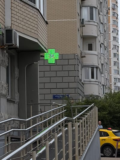 A bright green, pixelated cross sign with a caduceus symbol in the center is mounted on the side of a multi-story brick building. Below the sign, a blue street sign reads "АТЛАСОВА УЛИЦА" (Atlasova Street). A set of metal handrails leads up a ramp. In the background, a yellow taxi cab is parked, and a man in a light blue shirt stands near it. The scene appears to be in a residential area of Moskovskiy, Russia.