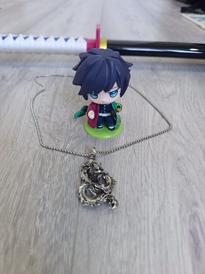 A silver dragon pendant necklace lies on a light gray surface. Behind it stands a small figurine of an anime character with dark blue hair and a green base. In the background, the hilt and part of the blade of a katana sword are visible. The hilt is wrapped in white, and there is a pink accent. The blade is shiny black.