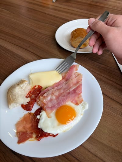A high-angle, close-up photograph displays a breakfast setting on a dark brown wooden table, likely indoors in Villajoyosa/la Vila Joiosa, Spain, though the precise location within the city is not visually discernible. In the foreground, a white, round plate holds a variety of food items: a fried egg with a bright orange yolk and set white, several strips of raw or lightly cooked bacon, a rectangular slice of yellow cheese, multiple slices of reddish cured sausage (possibly chorizo), and several thin slices of reddish cured ham (possibly jamón). A torn piece of white bread is also present on this plate. A human hand, with visible knuckles, holds a silver metal fork, with its tines positioned above the cheese and bacon on the foreground plate, indicating an active or impending eating action. In the blurred background, a second white, round plate contains a single golden-brown pastry, resembling a croissant.