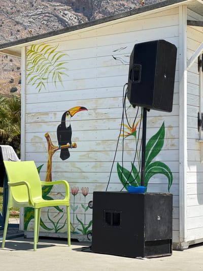 The image depicts an outdoor scene in the city of Capaci, Italy. In the foreground, a bright lime green plastic chair is positioned on a paved surface. Behind the chair, a white wooden structure with painted tropical motifs, including palm leaves, a toucan, and flowers, serves as a backdrop.
Mounted on a stand to the right of the chair is a large black speaker. Below this speaker, another black speaker cabinet rests on the ground. Several black electrical cables are visible, connecting the speakers. A small blue bowl is placed on top of the speaker cabinet.
In the background, a rocky mountain slopes downwards, and some greenery, likely palm trees, is visible to the left. The overall setting suggests a casual outdoor venue, possibly a beach bar or a small entertainment area. No people are present in the frame. The lighting indicates it is a sunny day.