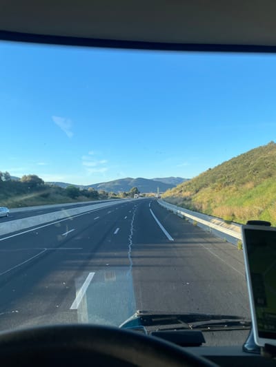 The image presents a first-person view from inside a vehicle, likely a truck, traveling on a multi-lane asphalt highway near Tresserre, France. The road ahead curves slightly to the left, marked with white dashed and solid lane lines, and shows a distinct crack running through the current lane's asphalt. To the left, a concrete barrier separates the highway from a parallel road where a light-colored sedan is visible, followed by a landscape of green vegetation, trees, and distant hills under a clear blue sky. To the right, a steep embankment covered in green and dry vegetation rises, bordered by a concrete safety barrier. Further down the highway, a white truck and other vehicles are discernable, along with a metallic power line pylon on the right side. The scene is illuminated by direct sunlight, casting long shadows across the roadway. In the immediate foreground, the top rim of a steering wheel is visible, and on the lower right, a dashboard-mounted navigation device displays a green map interface. The vehicle's windshield wipers are present at the bottom of the viewable area.