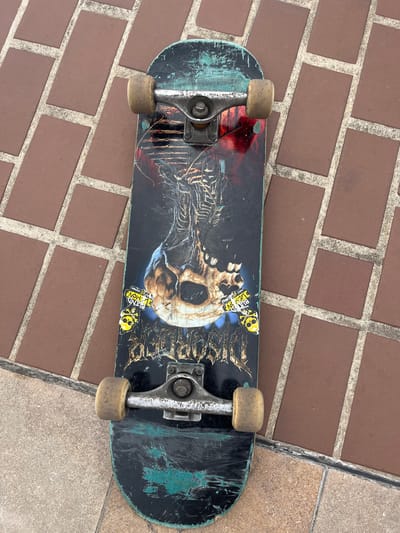 A black skateboard with a graphic of a skeleton and skull is lying on a brick-patterned sidewalk. The skateboard has two silver trucks and four yellowed wheels. The graphic on the skateboard features a skeletal ribcage with a skull at the center. There are also some stickers on the skateboard. The sidewalk is made of rectangular red bricks with white grout lines. The scene is outdoors, and the lighting suggests it is daytime. There are no people visible in the image. The skateboard appears worn, with scuff marks and faded paint, indicating it has been used. The overall environment suggests a casual urban setting. The location is the city of Créteil, France.