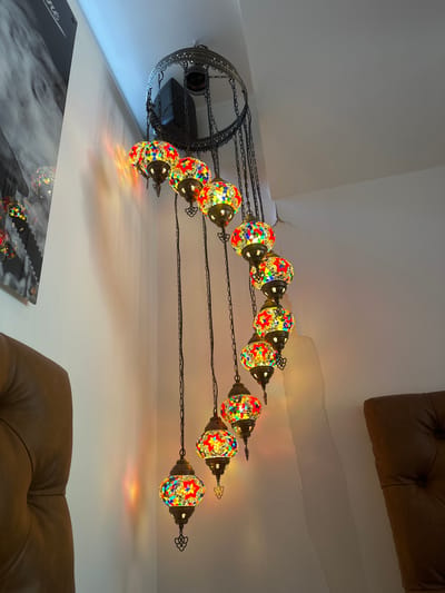 An illuminated and ornate mosaic chandelier dominates the upper portion of an indoor scene, likely in Haarlem, Netherlands. The chandelier, possibly of Turkish or Moroccan design, features a dark, intricately patterned metal frame from which multiple lamps are suspended by chains. Each lamp is spherical, crafted from numerous small, colorful mosaic glass pieces that glow brightly, casting warm, dappled light and vibrant reflections onto the white walls and ceiling. The lamps are arranged in an arcing pattern, adding to the chandelier's visual appeal.
The background consists primarily of plain white walls and a ceiling, which exhibits some minor cracks and imperfections, particularly visible towards the top right. In the upper right and lower right sections of the image, parts of a dark brown, tufted upholstered sofa or armchair are visible, suggesting a comfortable seating area. On the bottom left, a corner of a dark, glossy piece of art or a sign is visible, featuring an abstract, smoky design. The partial word "ne" can be deciphered on this item. The overall impression is of a warmly lit interior space, possibly a restaurant, cafe, or lounge, with a distinctive decorative centerpiece.