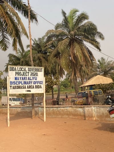 A sign reads "BIDA LOCAL GOVERNMENT PROJECT 2020. MAYAKI ALIYU DISCIPLINARY OFFICE TAKONASA AREA BIDA" with an arrow. It stands near palm trees and a roadside area. Buses are parked behind a low concrete barrier. A few figures are visible in the background near the buses and vehicles are parked along the road. The ground is covered in sand.