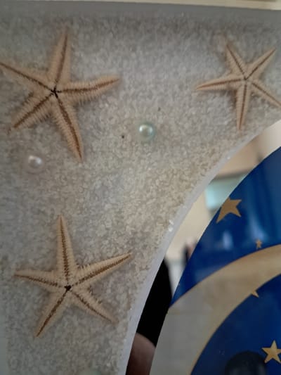 This close-up image captures a decorative arrangement featuring three light-colored starfish, two round pearls, and a bed of fine white granular material resembling sand. The objects are viewed through a clear, reflective surface, likely glass or a mirror.
One starfish is positioned in the upper left, another in the lower left, and a third in the lower right of the frame. A pale blue or light green pearl is visible near the center-left, and a white pearl is located in the lower-center. The textured surfaces of the starfish and the individual grains of sand are clearly discernible.
Reflected in the upper right portion of the clear surface is a vibrant blue object decorated with a yellow crescent moon and several yellow stars, suggesting a celestial or night sky theme. Below this reflection, a blurry, dark shape, possibly a person's arm or shoulder wearing dark clothing, is also faintly visible, indicating the indoor environment where this decoration is situated.
No specific activity or event is taking place; the image portrays a static decorative display. The overall aesthetic evokes a calm, beach-themed or celestial atmosphere. There is no visible text in the image. The location context for this media file is Gavar, Armenia.