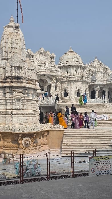 A large white temple with intricate carvings fills the frame. People ascend the steps towards the entrance. The structure features multiple spires and domes. A sign with writing is visible to the side of the steps. The sky is clear and pale blue. The architectural details of the temple are prominent, showcasing its ornate design.