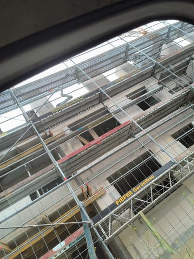 This is a photo taken from inside a vehicle, looking up and out at a construction site. The view is framed by a dark, curved object, likely the interior of the vehicle's window or frame.
The construction site features extensive scaffolding, made of metal poles and mesh platforms. The scaffolding surrounds a building under construction, which appears to be made of concrete blocks. Several floors of the building are visible, with open window frames and some exposed structural elements.
Yellow and red bands are visible on some of the scaffolding platforms, possibly indicating safety warnings or company branding. The words "PIEPER Gerüstbau" are printed in yellow on black bands on several parts of the scaffolding, identifying the company responsible for its erection. A yellow construction crane is visible in the background.
The sky is overcast and bright, suggesting daylight. Small yellow wildflowers are growing in the foreground, outside the construction site fencing. The overall impression is of an active construction project. There are no people visible in the image. The location is identified as Hamm, Germany.