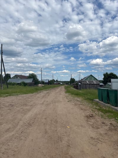 The image depicts a rural, unpaved dirt road in a village, likely in Russia, given the presence of wooden houses and the architecture. The road stretches into the distance, flanked by grassy verges and several rustic wooden structures. The houses appear to be single-story with weathered wooden siding and metal roofs. In the foreground on the right side of the road, there are three large green waste bins situated on a concrete base. The sky is partly cloudy, with dramatic white cumulus clouds against a bright blue background. Telephone poles and overhead wires are visible along the left side of the road, as well as further down the road, indicating utility infrastructure. The environment is one of a quiet, rural settlement with typical village housing and a dirt road, possibly in the city of Kurgan, Russia, judging by the context provided. There are no people visible in the image.