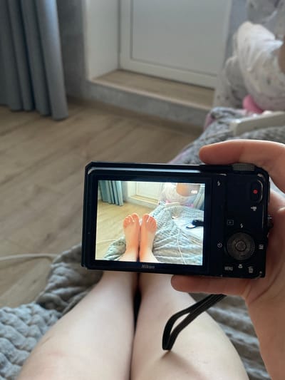 A person holds a Nikon digital camera, displaying an image on its screen. The image on the screen shows a close-up view of bare feet resting on a gray-and-white patterned bedspread. In the background of the image on the screen are a white wall and a stack of gray books. The location is Зубово, Russia.
