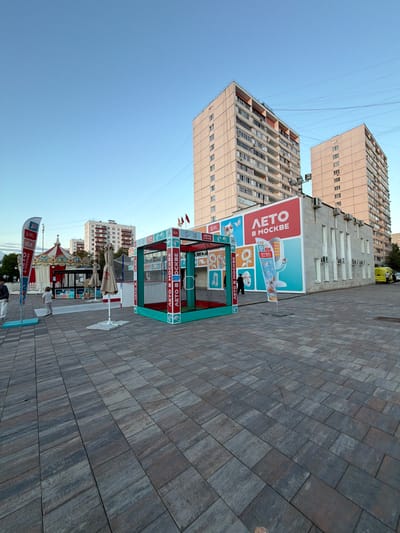 This image captures an outdoor scene in Moskovskiy, Russia. The setting features a wide, paved walkway with a repeating brick pattern, likely a public square or pedestrian area. Dominating the background are two tall, apartment buildings with numerous windows and visible air conditioning units. The sky above is a clear, pale blue, suggesting daytime with fair weather.
In the foreground, a small, enclosed play area or kiosk with a green and teal frame is set up. It has a net enclosure and is decorated with banners and posters. One prominent banner displays text in Russian, which appears to say "ЛЕТО В МОСКВЕ" (Summer in Moscow) in large red and white lettering, alongside illustrations of a cat, a sunflower, and a plane. Another smaller banner repeats the phrase "ЛЕТО В МОСКВЕ".
To the left of the play area, a carousel with colorful decorations is visible, hinting at a festive or recreational event taking place. Several flags are also seen attached to the play structure. A few people are present in the scene, including one person walking near the play area and others further in the background, suggesting a moderate level of activity. A yellow vehicle is parked in the background near one of the buildings. The overall impression is of a vibrant urban space during a summer event.