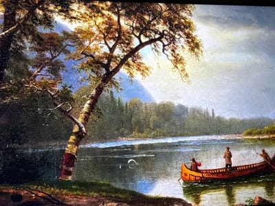 The image displays a photograph of a painted natural landscape, located within the city of Reus, Spain. The artwork depicts a wide river flowing through a valley, flanked by dense forests and distant blue mountains under a partially cloudy sky.

In the foreground on the left, a large deciduous tree with a gnarled, twisted trunk and exposed roots extends upwards and towards the right. Its foliage, rendered in hues of brown, gold, and green, suggests an autumn or late summer setting, with brighter light illuminating its upper branches. The ground immediately beneath the tree is dark, featuring patches of green and reddish earth.

The middle ground is dominated by the river, characterized by dark, reflective water showing ripples and currents. A distinct white, crescent-shaped disturbance is visible on the water's surface in the left-center, suggesting movement or a splash.

On the right side of the river, a reddish-orange canoe with visible wooden ribbing contains three individuals. The central figure stands, facing towards the right, actively engaged in fishing with a long, bent rod, indicating a catch or resistance. This individual wears earth-toned attire and a hat. In the bow, another person is seated or kneeling, wearing a reddish top. At the stern, a third person is seated or standing, facing right, holding a paddle to propel or steer the canoe. Reflections of the canoe and its occupants are visible in the water beneath them.

The background features a continuous line of dark green, densely forested hills or mountains along the riverbanks. Beyond these, lighter blue-hued mountains rise against a sky that transitions from bright, almost white light in the upper right quadrant to a soft blue and then to a cloudier, light grey-blue towards the far right. A subtle band of light blue and pale yellow is visible in the sky above the distant mountains, beneath the bright light source.