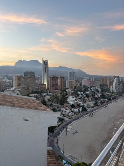 This is an exterior, high-angle, medium shot of the Benidorm skyline in Spain at sunset. The sky is a pale blue with streaks of pink and orange clouds. In the background, hazy mountains rise above a sprawling city of high-rise buildings, including the distinctive Torre Lúgano. A beach with light-colored sand and a gentle surf lines the foreground. A curved road separates the beach from the city, with palm trees and streetlights lining the promenade. Several cars are visible on the road. In the immediate foreground, a white, textured wall with a tiled roof angles into the frame from the left. There are no people clearly visible in the frame. The overall scene depicts a coastal city during twilight.