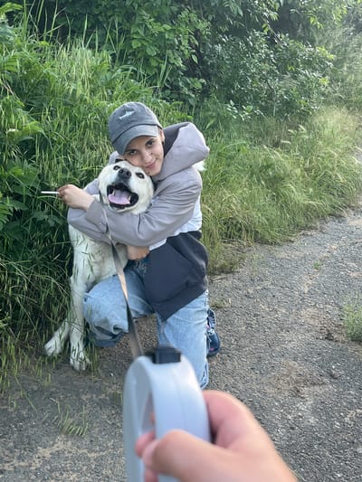 A young person kneels in a grassy area next to a paved path, embracing a large, cream-colored dog. The person wears a grey jacket, light-wash jeans, and a grey baseball cap. The dog has its mouth open. A retractable leash extends from the dog's collar, held by a hand in the foreground. The location is undetermined, it could not be determined from the visual information. There is a cigarette on the ground near the dog.