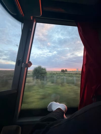 The image shows a view from inside a vehicle, likely a bus, looking out a window. The window is large and partially obscured by a red curtain on the right side. Outside the window, a blurry landscape is visible, suggesting the vehicle is in motion. The sky is filled with clouds, illuminated by the soft light of either dawn or dusk, with a hint of orange and pink near the horizon. In the distance, there appear to be some industrial structures. In the foreground, a person's legs and a hand wearing a white glove are visible, resting near the window. The overall impression is one of travel and observation of the changing light and landscape. The specific location within the city of Unknown, Romania cannot be determined from the visual information provided.
