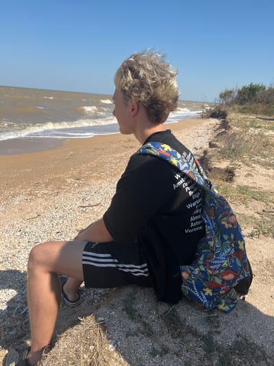 A young male with blond, curly hair sits on a sandy beach in Dolzhanskaya, Russia, looking out at the Sea of Azov. The sky is clear and blue. The waves are choppy, indicating a breezy day. The beach is covered in sand and shells. The male is wearing a black t-shirt with text printed on the back and black athletic shorts with white stripes. A backpack with a colorful floral pattern is slung over his right shoulder. His left leg is visible, showing a light scar on the shin. He is wearing black Crocs-style sandals. In the background, dry vegetation and sand dunes line the coast. The image captures a moment of quiet contemplation by the sea.