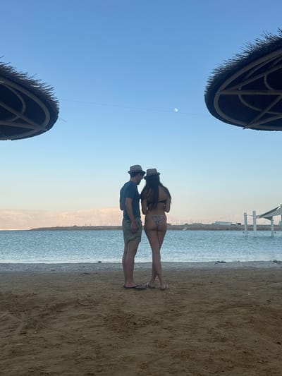 A man and a woman stand facing away from the camera on a sandy beach, looking out at a body of water. The man is wearing a blue t-shirt, khaki shorts, and a hat. The woman is wearing a bikini with a tiger-striped pattern and a hat. The water is calm and reflects the clear blue sky. In the distance, there are low, barren hills. Two thatched umbrellas provide shade on the beach. The overall scene suggests a relaxed vacation setting.