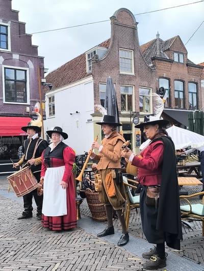The image depicts an outdoor street scene in the city of Alkmaar, Netherlands, under an overcast sky. Four individuals, dressed in historical 17th-century European costumes, are positioned in a line, facing slightly right, on a herringbone-patterned cobblestone street.
From left to right:
1. A man in a black tunic with white collar and cuffs, black breeches, and a black hat with a yellow feather, is playing a large brown drum with two sticks. His footwear consists of black ankle boots.
2. A woman wears a red dress with a black bodice, a white apron, a white head covering, and a wide-brimmed black hat. She holds a tambourine in her right hand. Her shoes are dark brown.
3. A man in a light brown tunic, breeches, white collar, and a wide-brimmed black hat, plays a long woodwind instrument (possibly a shawm or dulcian) with both hands. A small tan pouch with a dark cord hangs from his right hip. He wears black shoes.
4. A man in a dark red tunic, dark breeches, a black cape draped over his left shoulder, and a wide-brimmed black hat adorned with a large white feather, plays a stringed instrument resembling a lute or mandolin. A decorative brown leather pouch with a silver buckle is attached to his belt. He wears black shoes with ornate gold-colored buckles.
The background features historic Dutch architecture. On the far left, a dark brick building with a stepped gable shows a "TE KOOP ROGIER MAKELAAR" (For Sale) sign with a phone number (072-5021705) in a ground-floor window. A red awning covers a storefront with "AMSTEL BIER" and "HET HOOFD" visible. Adjacent to it, a white building facade has a street sign reading "GULPENWEG" and a mounted security camera. To the right, a prominent brick building with an ornate bell-gable displays a "GUINNESS" sign and an Irish flag in a window. In the mid-ground, multiple outdoor cafe tables with light-colored chairs and cushions are visible, along with a large black market umbrella on the left and a striped white umbrella on the right. A wicker basket sits on the ground next to the third musician. The architectural styles, business names, and visible street sign confirm the location as Alkmaar, Netherlands.