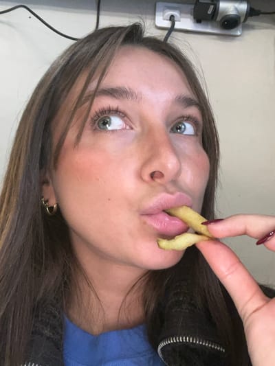 A close-up, chest-up indoor portrait depicts a young adult female in Aix-en-Provence, France. She has long brown hair, light green eyes directed upwards and to the right, and visible blush on her cheeks. Her lips are pursed, with two light green/yellow, fry-shaped snacks partially inserted into her mouth. Her right hand, exhibiting dark red nail polish, holds the snacks. She wears small gold hoop earrings in her left ear and a blue garment under a black zippered jacket. The background features an off-white wall, displaying a white power strip with a black cable and a black and grey electrical adapter plugged into its outlets, along with another loose black cable.