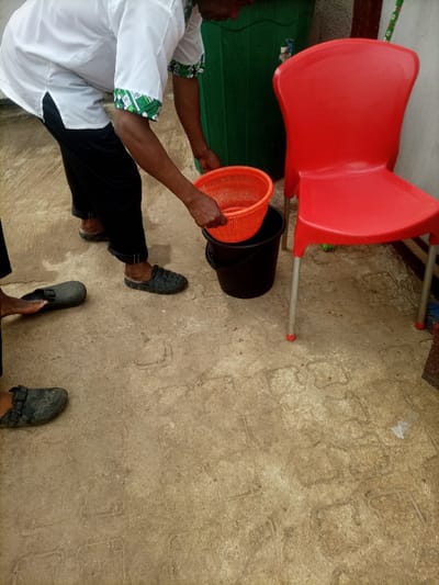 A person wearing a white shirt and black pants places an orange basket inside a black bucket. A red plastic chair stands to the right of the bucket. Behind the chair is a tall, green container. The person is wearing dark-colored sandals. Another person is visible in the lower left corner, also wearing dark-colored sandals. The ground is a textured, light brown color with imprinted designs.