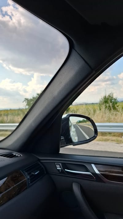 The image shows the interior of a car looking out the passenger side window. The foreground is dominated by the car's interior, including a portion of the dashboard with a wood-grain accent, an air vent, and a door panel with a chrome handle and lock buttons. In the middle ground, a side-view mirror reflects a road with white lines, suggesting the car is in motion. Outside the car, a rural landscape is visible, featuring a road with a metal guardrail, fields of dry grass or wheat, and some trees. The sky is partly cloudy with white clouds against a blue sky. The scene is likely captured on a sunny day, as indicated by the lighting and reflections. The location is specified as the city of Malo Konare, Bulgaria.