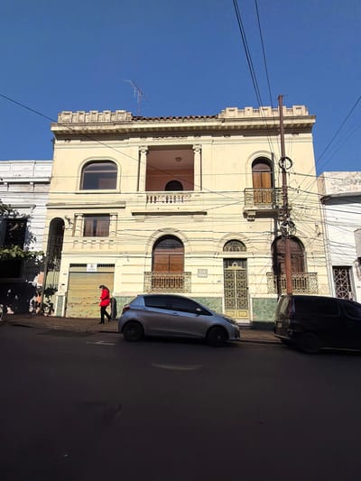 This is a photograph of a street scene in Asuncion, Paraguay, likely on a clear day with a bright blue sky. In the center of the frame is a multi-story, pale yellow building with ornate architectural details, including arched windows, balconies with decorative ironwork, and a crenellated parapet. The building's facade features classical elements like columns and decorative molding.

In front of the building, a silver hatchback car is parked on the street. To the right of this car, a dark-colored van is also parked. On the sidewalk in front of the building, a person wearing a red jacket and dark pants is walking from left to right. The building has a rolling metal garage door on the ground floor, partially open, and a decorative metal gate next to it. A sign with the text "ISLA FRANCIA" is visible on the building.

The street in the foreground is paved and appears to be in shadow in some areas. A utility pole with numerous wires is prominent in front of the building. The overall impression is of an urban street with older architecture.