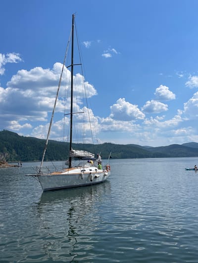 A sailboat is moored on a calm lake. The boat is white with wooden accents and has a tall mast. Two people are visible on the deck of the boat, and one person is on a paddleboard in the distance. The sky is blue with scattered white clouds. Lush green hills rise from the far side of the lake, suggesting a mountainous or hilly terrain. The water is slightly rippled, reflecting the sky and surrounding landscape. The lighting suggests it is daytime, likely late morning or afternoon, under clear, sunny weather. The scene evokes a sense of peaceful leisure and outdoor recreation on the water. The location is Divnogorsk, Russia. No visible text is present in the image.