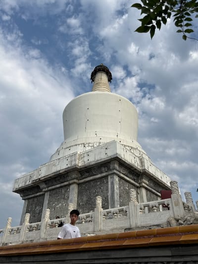 In Xicheng District, China, a young person in a white t-shirt stands on a decorative balustrade, looking towards the camera. Behind them, a large, white, stupa-like structure dominates the frame under a cloudy sky. The stupa features a tiered base with grey brickwork and ornate white stone railings with carved posts. Green leaves hang from the upper right corner of the image, partially obscuring the sky. The stupa itself is cracked in places, suggesting age or weathering. Other figures are visible in the background, blurred and less distinct.