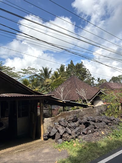 The image depicts a scene on a street in Susut, Indonesia. In the foreground, a pile of dark, irregular volcanic rocks sits on the side of a dirt road, bordered by green grass. Behind the rocks, a low concrete wall runs horizontally across the frame. Beyond the wall, traditional Indonesian houses with dark, tiled, pyramidal roofs are visible, nestled among lush green trees, including palm trees. Overhead, a tangle of black electrical wires stretches across the bright blue sky, which is dotted with fluffy white clouds. To the left of the houses, a partially visible structure with a corrugated metal roof and wooden supports suggests a simple building or shelter. The overall impression is one of a rural or semi-rural residential area, characterized by the natural landscape and the distinctive architecture of the region. There are no people or animals visible in the image.