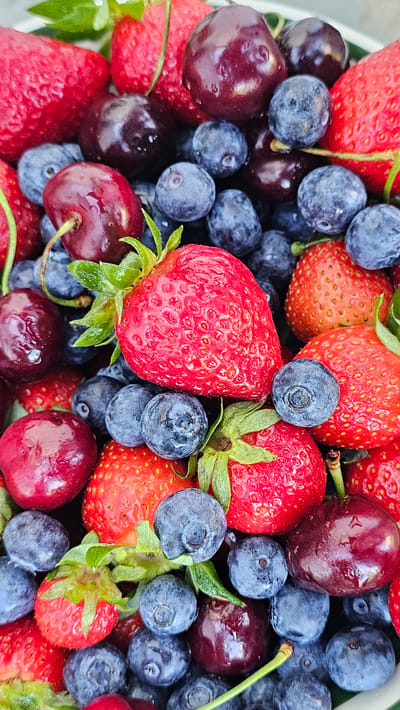 The image displays a close-up, overhead view of a dense assortment of mixed fresh berries, filling the entire frame. The primary subjects are numerous red strawberries, abundant deep blue to purplish blueberries, and several dark red cherries.
The strawberries vary in size, exhibiting textured surfaces with visible achenes and some retaining their bright green calyx and short stems. Many appear damp, with small water droplets visible. The blueberries are small, spherical, and uniformly distributed, displaying a natural powdery bloom and some moisture on their surfaces. The cherries are round, glossy, and dark maroon, also showing signs of wetness; a few have light green stems attached.
The berries are tightly packed together, suggesting they are contained within a vessel, though only minimal hints of a light-colored rim are visible at the very top and bottom edges of the frame. There are no people, actions, or specific environmental features depicted beyond the fruit itself. The lighting is bright and even, illuminating the natural colors and textures of the produce. The setting is undefined; no specific environmental or geographical details, such as the city of Slengiai, Lithuania, are discernible from the image.
