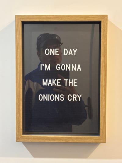 A framed message board is centered on a light-colored, plain wall. The frame is rectangular with a light brown, possibly wooden, finish. The board itself has a dark, reflective surface displaying white, uppercase text that reads: "ONE DAY I'M GONNA MAKE THE ONIONS CRY".
A reflection of a person is visible on the dark surface of the board, occupying the central portion of the frame. The reflected individual is holding a dark-colored smartphone, positioned as if taking the photograph. The person wears a dark baseball cap with sunglasses resting on its brim, a dark blue jacket with a white or light-colored horizontal stripe across the upper chest, and an earring in the visible left ear. A metallic ring is worn on a finger of the hand holding the phone. To the right within the reflection, a dark, horizontal door handle is visible, suggesting a door is present in the reflected background.
A faint shadow is cast by the bottom and right edges of the frame onto the light-colored wall, consistent with illumination from an overhead light source. No visual cues within the image identify a specific location within Asnières-sur-Seine, France; the scene depicts an interior space.