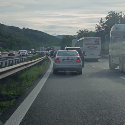 The image depicts a highway in Gorno Varshilo, Bulgaria, on a cloudy day. Multiple vehicles are on the road, including cars, a bus, and a truck. A silver Volkswagen sedan with license plate "CA 9573 HC" is in the foreground, facing away from the camera. Traffic appears to be moving slowly or stopped, suggesting a traffic jam. The highway is flanked by a grassy embankment and a metal guardrail. In the background, lush green hills rise up, covered in trees. The sky is overcast with visible clouds. The overall scene suggests a moment of congestion on a major road in a semi-rural or mountainous area.