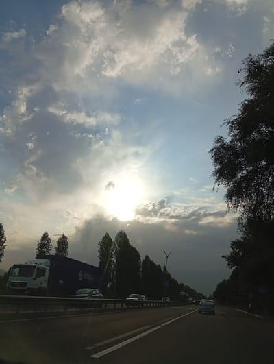 The image depicts a cloudy sky with the sun partially obscured by clouds, casting a bright glow in the upper center of the frame. The scene appears to be a highway in Uzbekistan, likely near Kara Kitay, given the context. On the left side of the highway, a large white truck with a blue shipping container is visible, bearing the "Atasu" logo. Several cars are driving on the highway in the same direction as the camera. The right side of the highway is lined with trees, some of which are silhouetted against the sky. The road itself has clear lane markings, and a metal guardrail separates the highway from the shoulder. The overall lighting suggests it might be late afternoon or early morning.