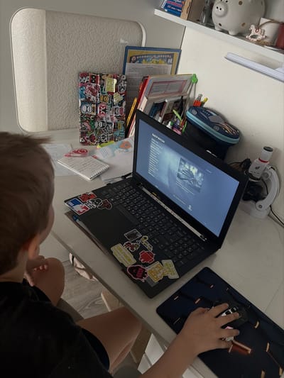 A young individual, viewed from behind, is seated at a white desk in what appears to be a study area within a residence in Novosaratovka, Russia. The person, with light-colored hair and wearing a dark t-shirt and shorts, is operating a black laptop computer. Their right hand is positioned on a white and black gaming mouse, which rests on a dark gaming mousepad with orange accents.
The laptop's screen displays a menu labeled "ВЫБОР МИССИИ" (MISSION SELECT) with an image of what appears to be a snowy, war-torn environment, indicating a video game is active. The laptop's palm rest and surrounding surfaces are extensively covered with various stickers, including designs related to "NBA," "Android," "Star Wars," and text such as "GIRLFRIEND" and "GOT MY WIFI FLY FOR EDI."
On the left side of the desk, next to the person, are several scattered papers and notebooks, including a spiral-bound notebook with handwritten entries and a red pen. Further left, a tablet or similar device encased in a heavily stickered cover stands upright. On the right side of the desk, behind the laptop, is a blue race car-shaped pencil case. A white microscope with red detailing is positioned on the far right.
Above the desk, a white shelf is visible, holding a white piggy bank with dark spots, a small rocking horse figurine, and an assortment of books and papers, including one featuring Russian text. The immediate background shows a light-colored wall or furniture, possibly a cabinet. The floor under the desk is light-colored.