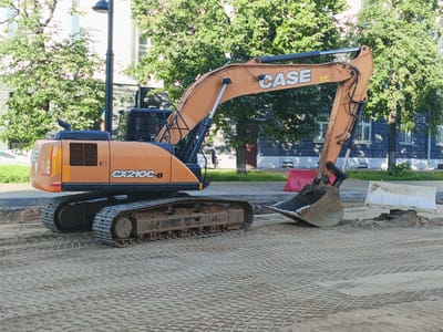 An orange CASE CX210C-8 excavator sits on a construction site in Yaroslavl, Russia. The excavator is positioned on a bed of sand that has a patterned grid imprinted on it from the movement of heavy machinery. The machine's tracks are caked with dirt, indicating recent use. To the left of the excavator, a paved walkway is visible, bordered by grass and trees. Behind the excavator, a building with light-colored walls and dark stone accents stands, with large trees providing shade. The building has several windows with white grates. Two red traffic barriers are placed in front of the building, and a white plastic barrier is further to the right, near a small mound of dirt. The scene suggests an ongoing construction or roadwork project. The sun is shining, casting shadows and indicating it is likely daytime, possibly mid-morning or afternoon, given the brightness of the light. No people are visible in the image. The text "CASE" is prominently displayed on the excavator's boom, and "CX210C-8" is visible on the machine's body. A triangular warning symbol with an exclamation mark is also present on the excavator's side.
