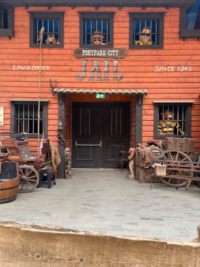 The image depicts the exterior of a wooden building with an orange facade, designed to resemble a jail. The building is located in the Ponypark City attraction in Collendoorn, Netherlands. Above the entrance doors, the words "PONYPARK CITY JAIL SINCE 1845" are visible, along with the inscription "LAW & ORDER" on the left side of the building. Three windows on the upper level of the building show cartoon figures, resembling prisoners, peering out from behind bars. The entrance features double wooden doors. In front of the building, to the left, is a horse-drawn carriage with a barrel and a wagon wheel. To the right, there are stacked wooden crates, saddles, a cowboy hat, and a wagon wheel. The ground is paved with bricks. The scene is outdoors, with clear daylight.