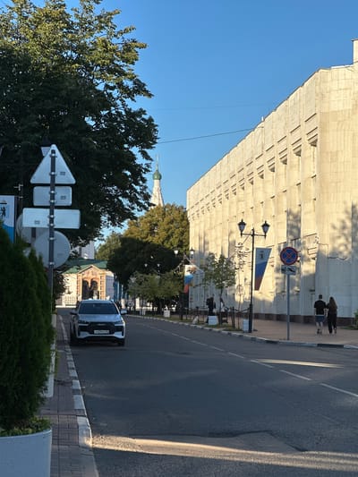 The image depicts a street scene in Yazvitsevo, Russia, bathed in the warm light of late afternoon. A modern silver car with a license plate starting with "H439 PC 76" is parked on the left side of the road, close to a sidewalk lined with manicured green bushes. The road itself is paved and has visible lane markings.
To the left, a traffic signpost with multiple triangular and rectangular signs stands prominently. Beyond the car, a building with an arched entrance and decorative facade is visible, with a Russian flag banner hanging to its right. Further in the background, the top of a white church tower with a green dome and cross pierces the clear blue sky.
On the right side of the street, a long, pale-colored building with a textured facade stretches into the distance. This building features repetitive vertical indentations and windows. A lamppost with two ornate lamps stands in front of it. Two people, a man and a woman, are walking away from the camera on the sidewalk in front of this building. Another lamppost is visible further down the street. The overall environment suggests a well-maintained urban area with architectural elements hinting at both historical and modern influences.