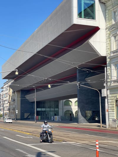 In Bratislava, Slovakia, a person rides a motorcycle on a street with tram tracks. The motorcycle is a classic cruiser style with a black finish and chrome accents. The rider is wearing a light blue shirt, dark pants, and a black helmet. In the background, a modern building with a distinctive cantilevered design dominates the scene. The building features a gray, perforated metal facade and bright red structural elements visible underneath. Adjacent to this modern structure is an older, light-colored building with classical architectural details. The sky is clear and blue, indicating a sunny day. The street has multiple lanes, a red-painted lane, and yellow markings. There are also overhead wires and streetlights present. A white car is seen in the distance, driving away from the camera.