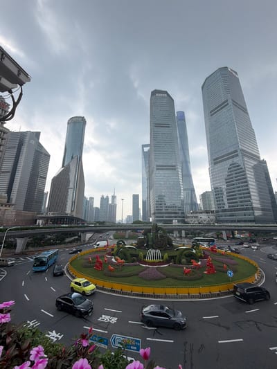 The image depicts a wide-angle view of a busy intersection in Pudong, China, dominated by modern skyscrapers. In the foreground, a circular roundabout features elaborate floral arrangements and decorative red horse sculptures. Several vehicles, including a blue bus, a yellow taxi, and black cars, are navigating the roundabout. Overhead, an elevated roadway with traffic can be seen. The background is filled with towering glass-fronted skyscrapers, indicative of a major urban center. The sky is overcast, suggesting cloudy weather. The overall scene captures the dynamic urban landscape of Pudong, characterized by its impressive architecture and active street life.