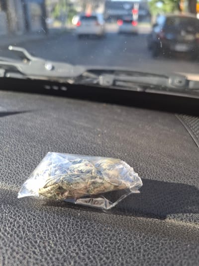 The image shows a clear plastic bag containing a green substance, likely plant matter, sitting on a textured dark surface, possibly a car dashboard. A windshield with wiper mechanisms is visible in the background, reflecting a blurred cityscape with buildings, traffic lights, and vehicles. The reflection suggests the photo was taken inside a vehicle. It's impossible to confirm that the picture was taken in Sarandí, Argentina, as there's no identifying information in the image, but the request specified that location.