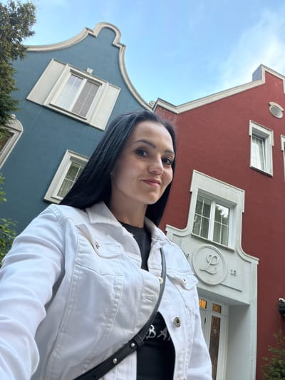 A woman with long black hair and fair skin, wearing a white denim jacket over a black t-shirt, stands in front of two buildings. The building on the left is painted teal and features a curved gable with a white trim and a single window. The building on the right is painted red and has a more angular roofline, also with white trim, and multiple windows. The woman is looking towards the camera, with her face turned slightly to her right. She is wearing makeup and has freckles on her nose and cheeks. A black bag with silver studs is slung across her body. The sky above is a light blue with a few wispy clouds. The architectural style of the buildings, particularly the gables and decorative elements, suggests they are located in Svetlogorsk, Russia, a resort town known for its distinctive architecture.